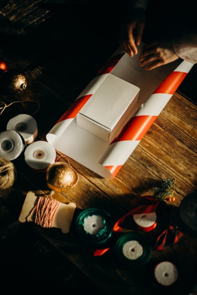 Hands wrapping a Christmas present with red and white paper, surrounded by festive decorations.