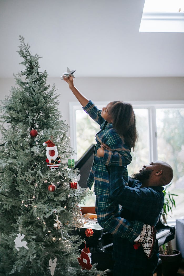 Father lifts daughter to place star on Christmas tree, celebrating holiday spirit indoors.